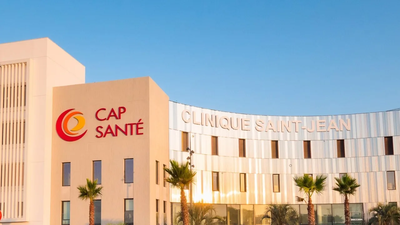 Façade crème d'un bâtiment à partie courbe en lames métalliques, palmiers devant, sous un ciel bleu