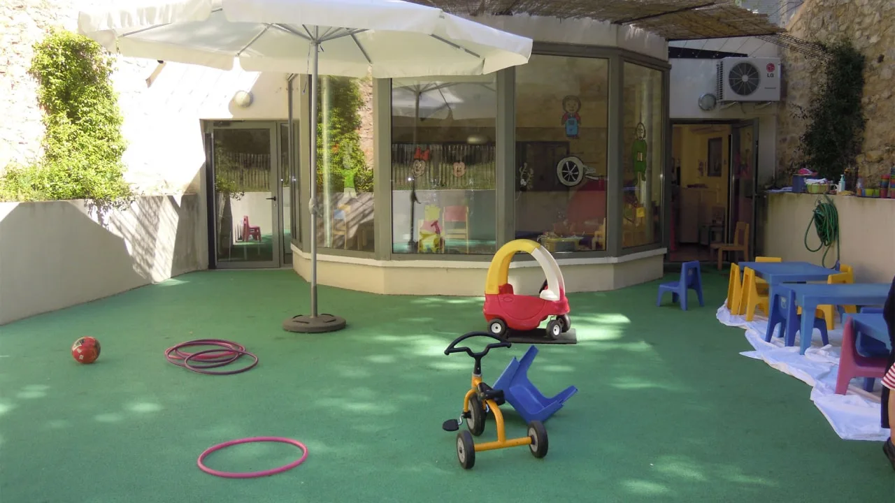 Espace de jeux extérieur à sol vert, avec un parasol, une voiture en plastique rouge et un tricycle jaune