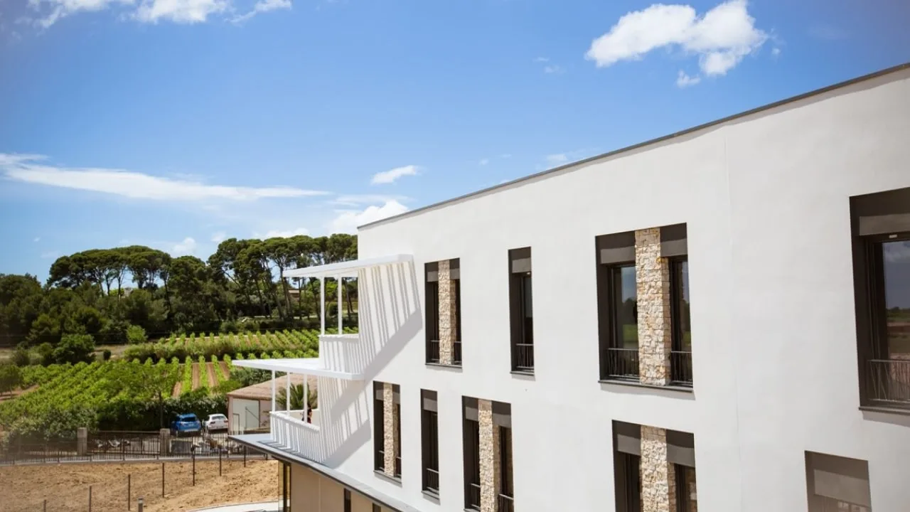 Angle d'une façade blanche avec structure à claire-voie, vignes et pins au fond sous un ciel bleu