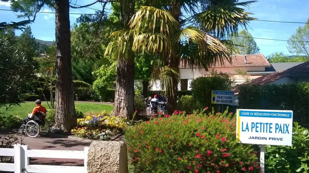 Jardin fleuri avec palmiers, une personne en fauteuil roulant sur une allée et des panneaux bleus et blancs