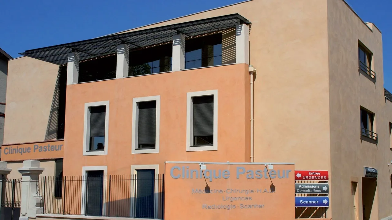 Façade orangée surmontée d'une structure métallique à lames, et panneaux de direction vers les urgences et le scanner