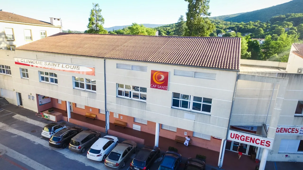 Vue en plongée d'un bâtiment crème à toit de tuiles, voitures garées devant et panneau Urgences rouge et blanc