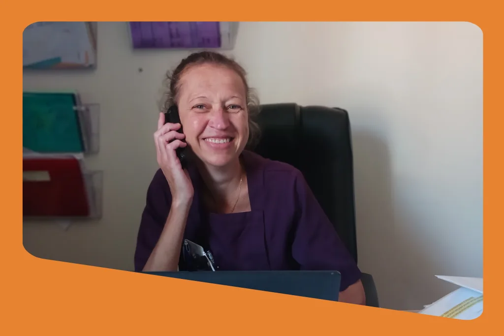 Femme souriante, téléphone à l'oreille, haut violet, assise dans un fauteuil noir devant des pochettes murales colorées